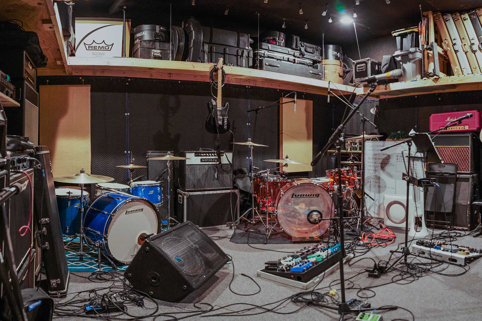 A large rehearsal room at Sonance Rehearsal Studios Brown Lane location with multiple drum sets, microphones, guitars, amplifiers, and various musical equipment scattered throughout the room.