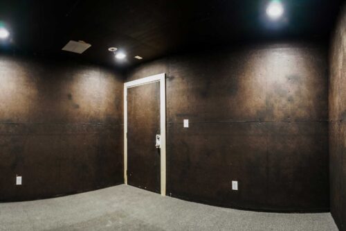 An empty rehearsal room at Sonance Rehearsal Studios Brown Lane location with black walls, bright overhead lighting, and a carpeted floor.