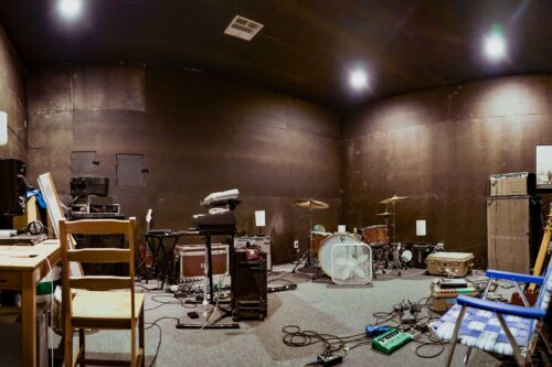 A large music rehearsal room at Sonance Rehearsal Studios Brown Lane location with instruments, amplifiers, cables, chairs, and various equipment scattered around. The room has dark walls and some overhead lights.