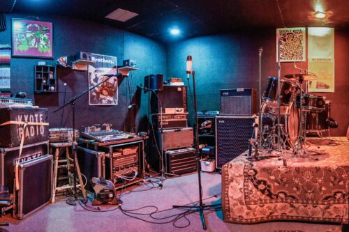 A colorful music rehearsal room at Sonance Rehearsal Studios Brown Lane location with various instruments, including drums, amplifiers, microphones, and sound equipment. Walls are adorned with posters, and a rug covers a drum riser.