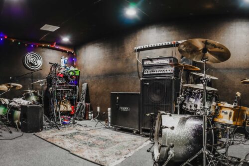 A music rehearsal room at Sonance Rehearsal Studios Brown Lane location with various instruments, including two drum kits and electronic equipment, set up against dark walls. Multicolored lights and cables are seen in the background.