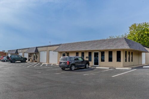 The Sonance Rehearsal Studios Reinli Street location, comprised of beige commercial buildings of rehearsal rooms with large windows and garage doors and a parking lot with several cars parked outside.