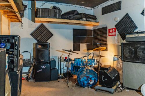 A room at Sonance Rehearsal Studios Reinli Street location with a blue drum kit, a large PA speaker, and a black amplifier. Acoustic foam panels cover parts of the walls, and a wooden shelf holds various cases and equipment.