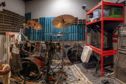 A music rehearsal room at Sonance Rehearsal Studios Reinli Street location with a drum set, cymbals, electronic equipment, soundproofing panels on the walls, and various pieces of musical and sound equipment scattered around.