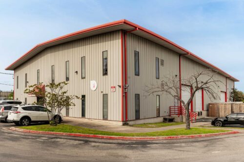 The Sonance Rehearsal Studios West Powell Lane location, a two-story industrial building of rehearsal rooms, surrounded by parked cars, trees, and loading docks with red railings.