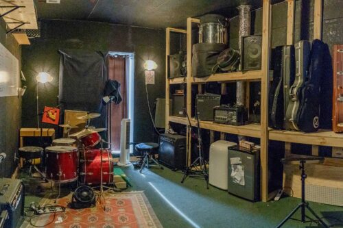 A music rehearsal room at Sonance Rehearsal Studios West Powell Lane location with a drum set, amplifiers, guitar cases, and various pieces of musical equipment organized on wooden shelves.