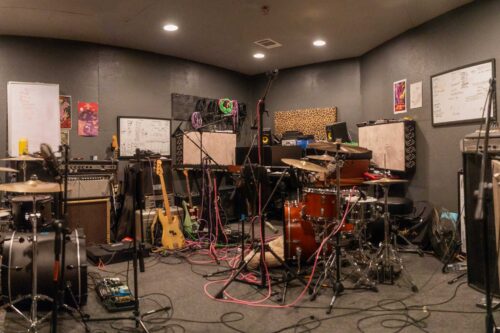 A music rehearsal room at Sonance Rehearsal Studios West Powell Lane location with various instruments including drums, guitars, amplifiers, microphones, and numerous cables scattered around.