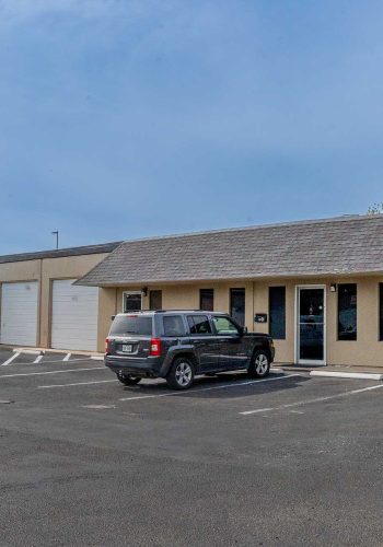 The Sonance Rehearsal Studios Reinli Street location, comprised of beige commercial buildings of rehearsal rooms with large windows and garage doors and a parking lot with several cars parked outside.