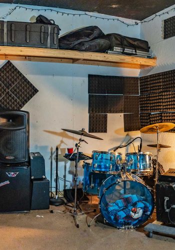 A room at Sonance Rehearsal Studios Reinli Street location with a blue drum kit, a large PA speaker, and a black amplifier. Acoustic foam panels cover parts of the walls, and a wooden shelf holds various cases and equipment.